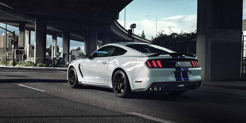 A white with blue racing stripes 2019 Ford Mustang driving under a city overpass. - Dann's Discount Auto Sales Visalia, CA